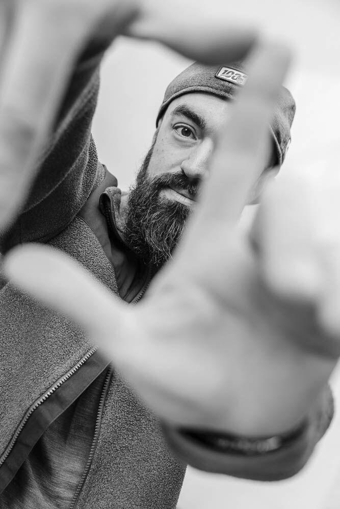 Schwarzweiß-Portrait von Fotograf Marvin Rust mit Mütze, die Hand zur Kamera ausgestreckt